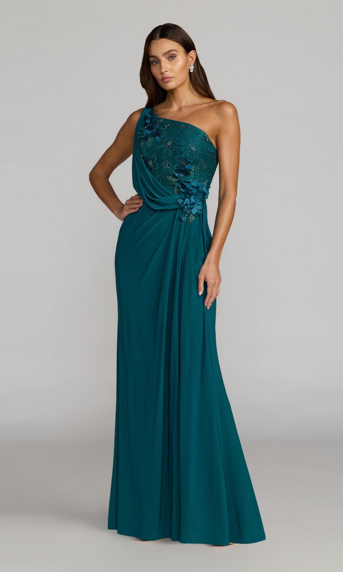 A woman stands against a plain light gray background, wearing the Mac Duggal 13210 long formal dress in Ocean—a one-shoulder, floor-length gown adorned with elegant draped accents and 3D flower detailing.